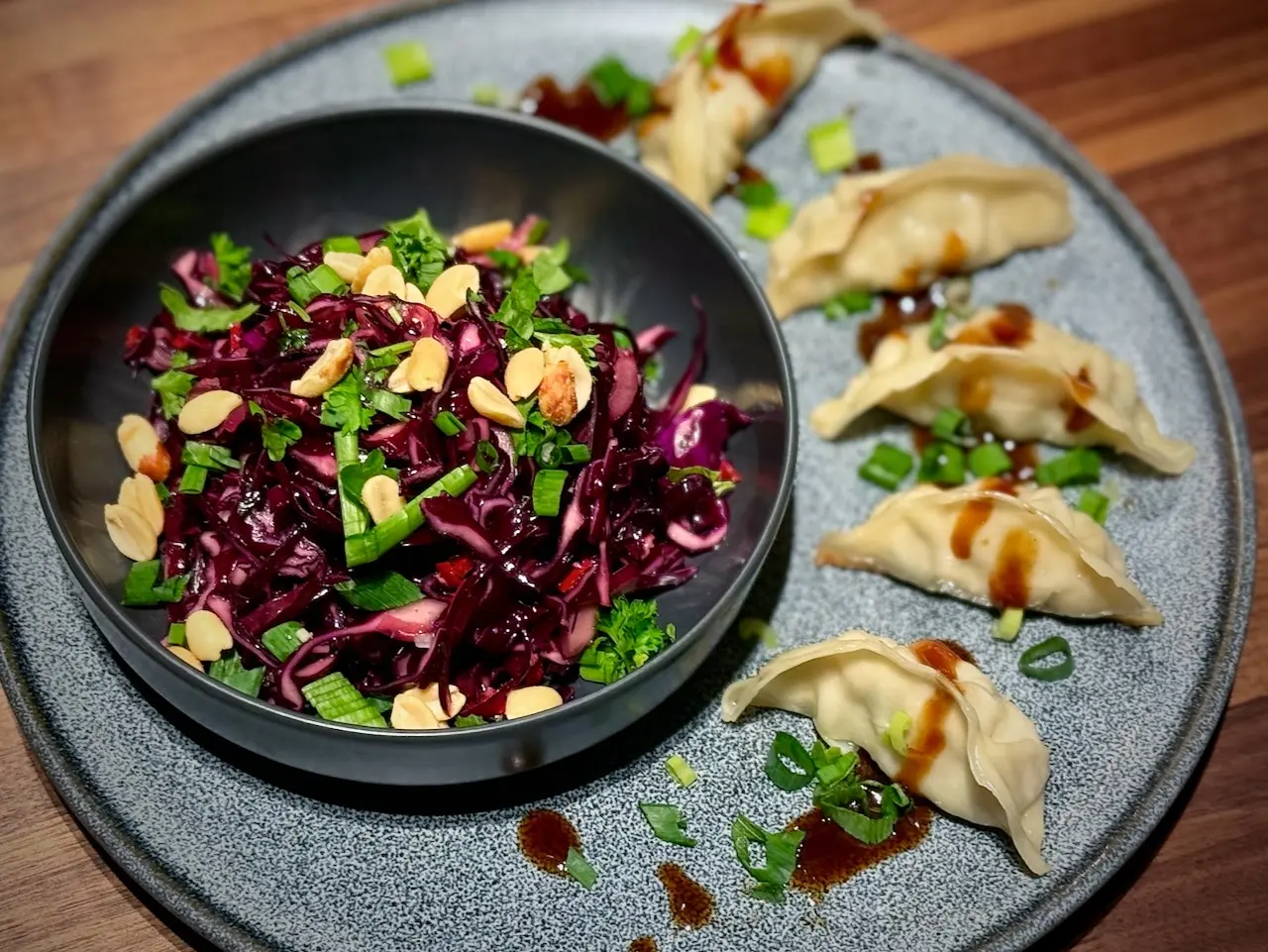 A dark bowl filled with vibrant purple Asian red cabbage slaw topped with peanuts and cilantro, served on a plate next to steamed dumplings.