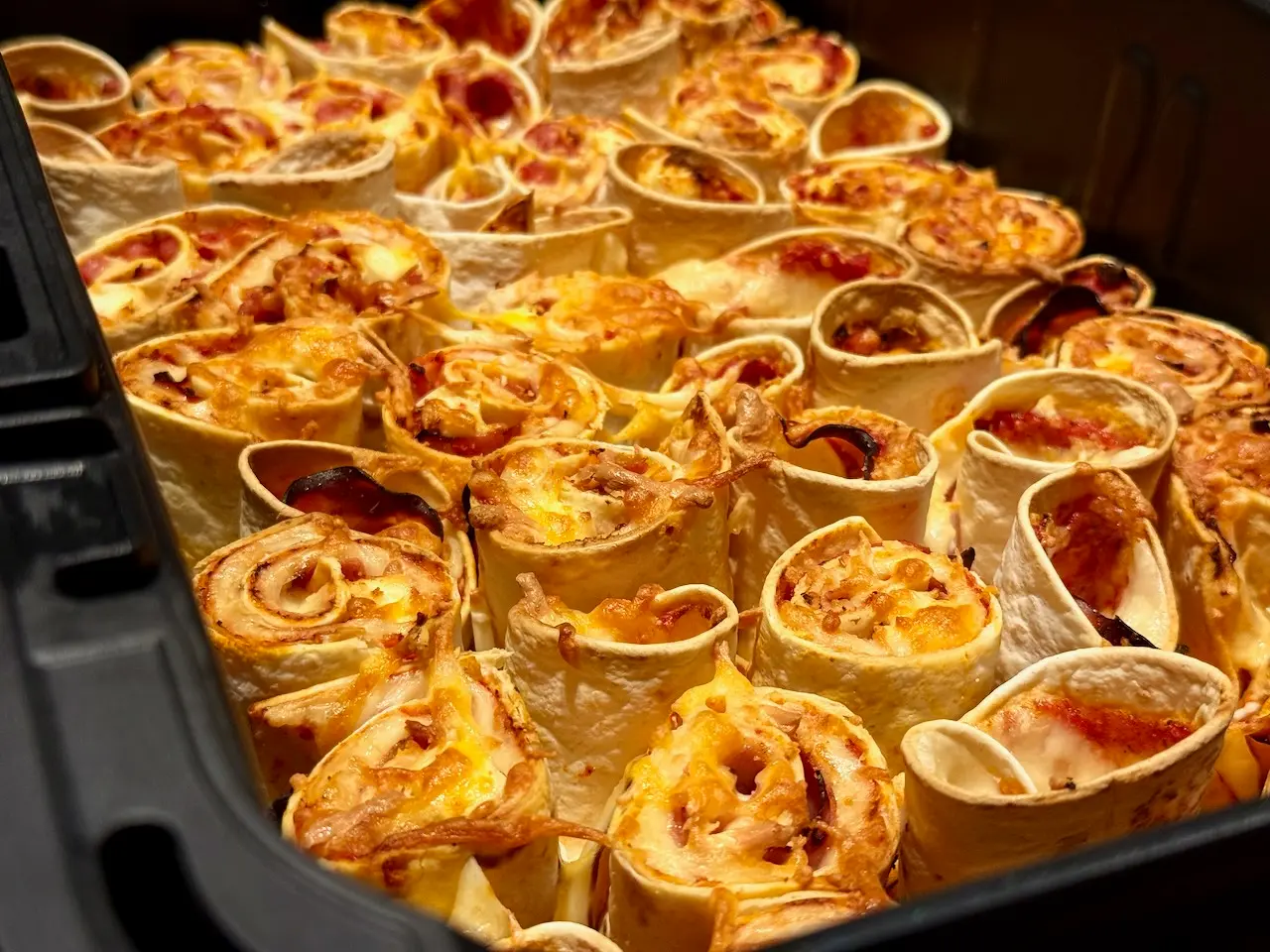 A basket full of golden brown air fryer tortilla pizza rolls filled with ham and cheese, topped with melted mozzarella.